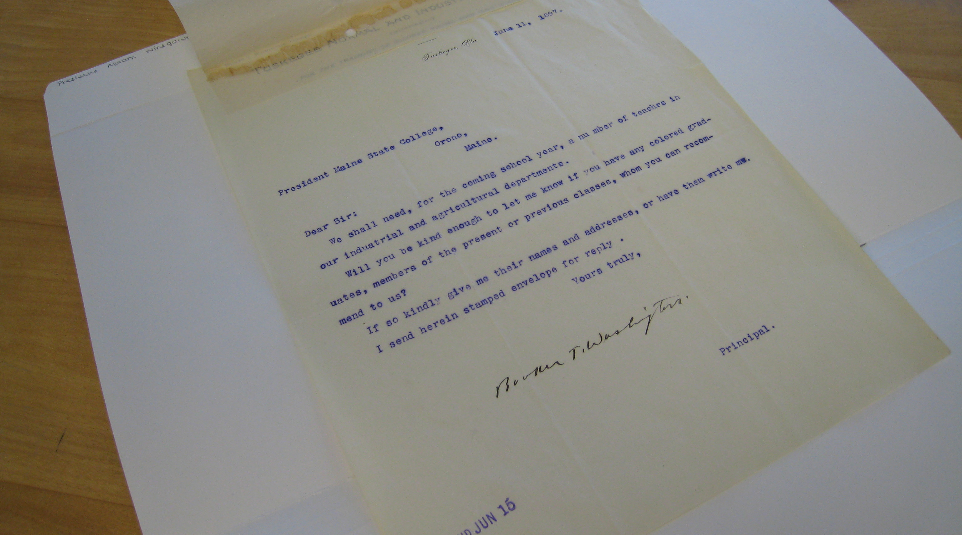 A close-up image of an old letter written on yellowed paper, typed with a typewriter. The top of the letter shows some damage, possibly from age, with a tear on the upper edge. The paper is lying flat on a surface, and a white envelope is partially visible in the background.