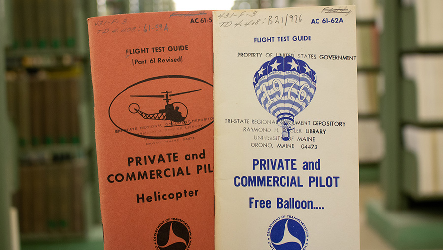 Two old flight test guide booklets are placed side by side on a table. The left booklet is titled "Private and Commercial Pilot Helicopter" with a small image of a helicopter, while the right one is titled "Private and Commercial Pilot Free Balloon..." with an image of a hot air balloon. Both booklets are marked with government property labels and a stamp indicating they belong to the Tri-State Regional Government Depository at the University of Maine. Bookshelves filled with other books are blurred in the background.