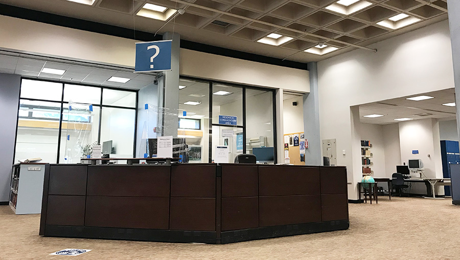 Information desk in a library with a blue question mark sign overhead, near bookshelves, a globe in a floor stand, printers, and computer stations.