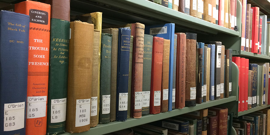 A close-up view of a library bookshelf filled with old and worn hardcover books. The books vary in color, size, and condition, with some spines showing faded or peeling covers. The books are neatly arranged on green metal shelves, with additional books visible in the background on higher shelves.