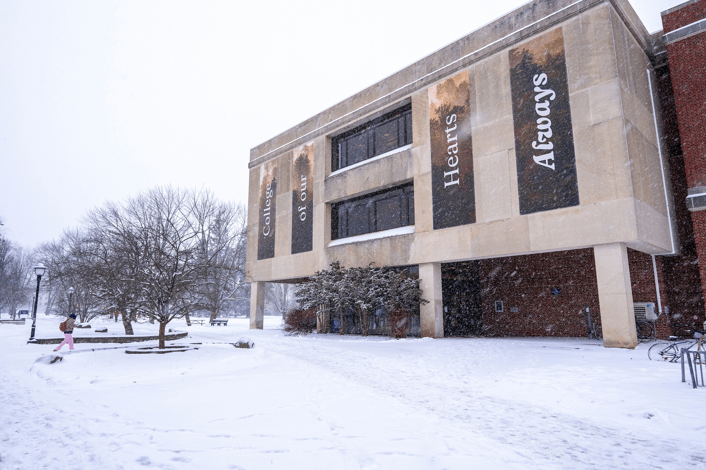 A large concrete and brick library building with banners displaying the words "College of our Hearts Always." Snow is falling, and the ground is covered in fresh snow. A person in a pink coat walks along a shoveled path, while leafless trees surround the building.