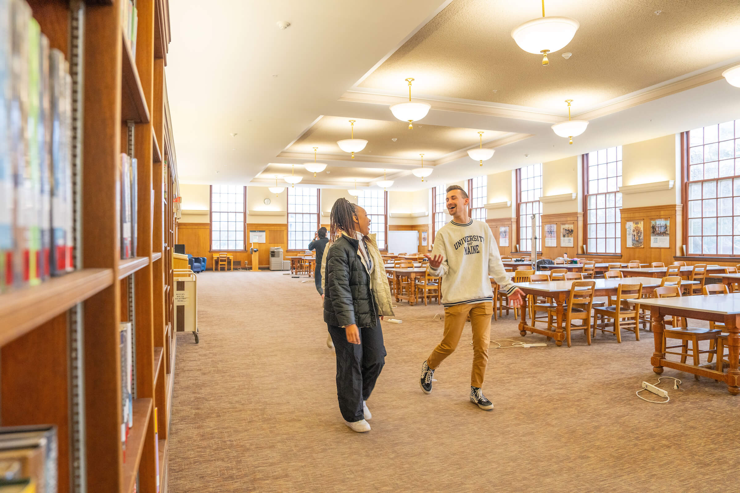 A spacious academic library with tall windows allowing natural light to fill the room. Wooden bookshelves line the left side, while large wooden tables and chairs are spread throughout. Two students are walking and talking with smiles on their faces.