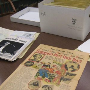 A table displaying archival materials, including vintage newspapers and magazines. The front page of a Detroit News article titled "Gardeners Must Raise Bugs" is prominently visible with colorful illustrations and photographs. Nearby, copies of a publication titled Independent Woman are stacked, and a white archival storage box labeled with handwritten notes contains folders of additional documents. The setting appears to be a library or archive reading room.