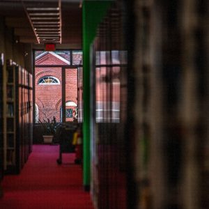 A long, dimly lit library hallway with red carpet leads to a large window at the end, through which a brick building with arched windows is visible. Bookshelves line both sides of the hallway, and a book cart is positioned near the window.