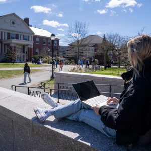 A student sits on a stone ledge outside on a sunny day, working on a laptop with legs crossed.