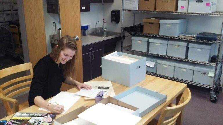Woman sitting at a table, smiling. A box and folders sit on the table. A shelf sits across from her, loaded with boxes.