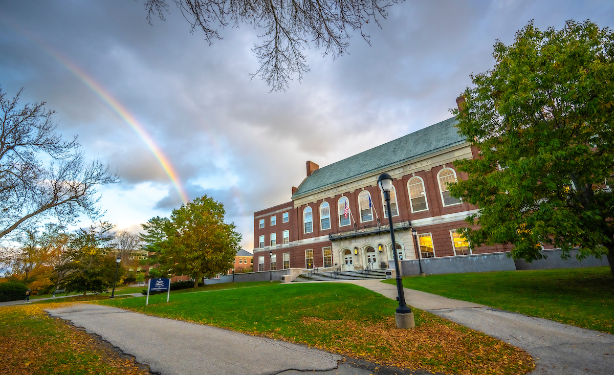 A red-brick building with a green roof, arched windows, and white steps is surrounded by green grass and trees with some autumn leaves. A rainbow is visible in the sky to the left of the building. Two paved paths lead towards the building, and a lamppost stands in the foreground.
