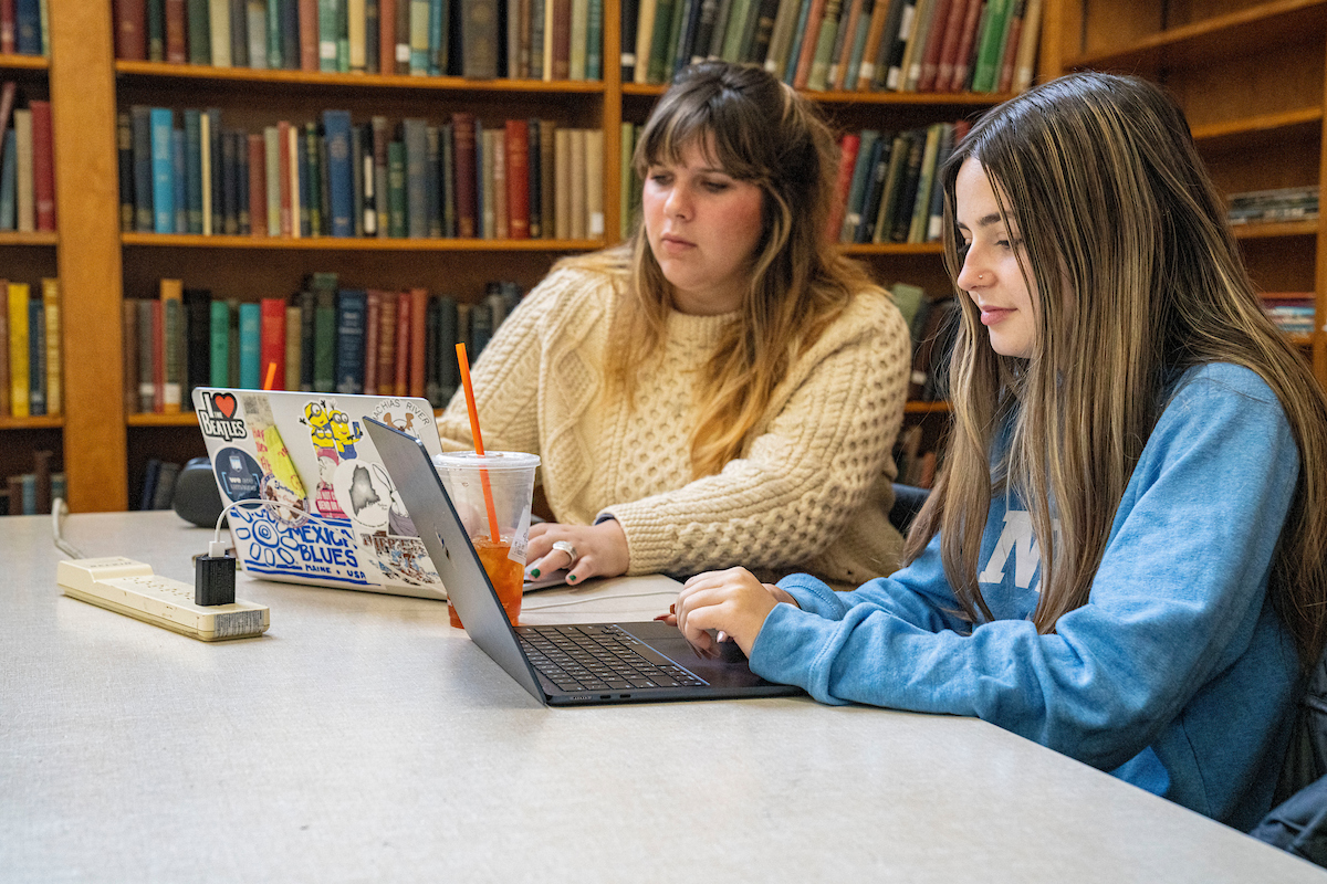 Two students sit at a table in what appears to be a library. The student on the left, with brown hair and wearing a cream-colored sweater, looks at a laptop with several stickers. Next to her laptop is a cup with a straw and a power strip. The student on the right, with long brown hair with blonde highlights and wearing a light blue sweatshirt, is actively typing on a black laptop. A wall of bookshelves filled with books is visible in the background.