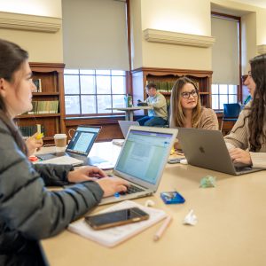 Three students sit around a large table in a library, each working on a laptop and engaging in conversation. Open notebooks, pencils, snacks, and phones are scattered on the table.