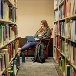 A student sits in a wooden chair at the end of a library aisle, working on a laptop and smiling. Bookshelves filled with colorful books line both sides of the carpeted aisle