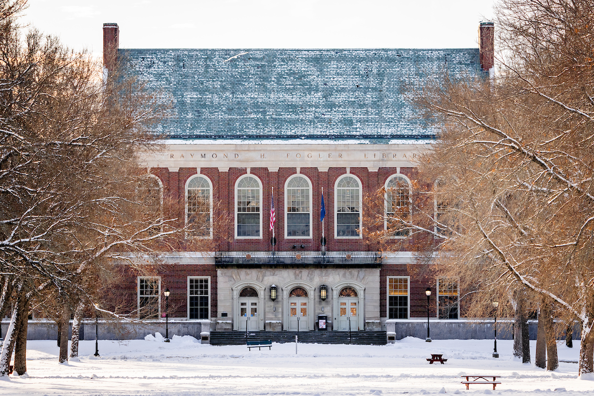Front view of Fogler Library in winter, with snow covering the ground and bare trees framing the red-brick building. The building features arched windows, a slate roof dusted with snow, and American and Maine flags above the main entrance.