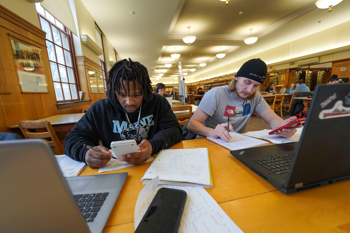 Two people study at a library table with laptops, notebooks, and calculators, surrounded by bookshelves and other students.