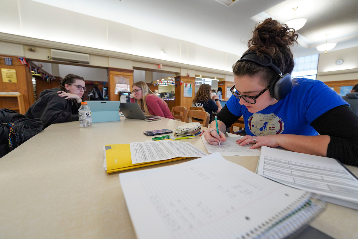 A group of people are seated at a table in a library or study area, engaged in studying and working on laptops. One person in the foreground, wearing headphones, is writing on paper with notebooks and documents spread out. 