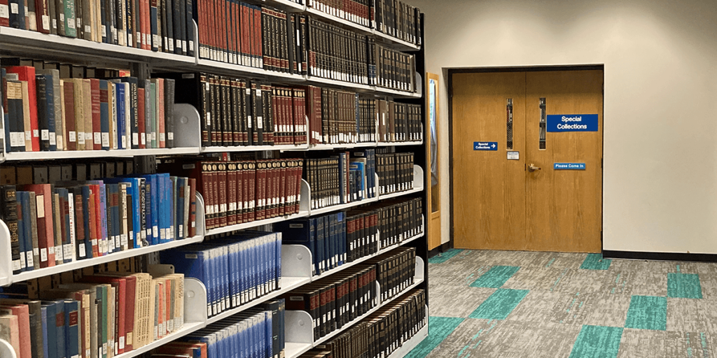 A library interior with several shelves filled with books on the left side and a set of closed double wooden doors on the right. The doors have blue signs reading "Special Collections" and "Please Come In." The floor is carpeted with a pattern of gray and teal squares. The overall atmosphere is quiet and organized.