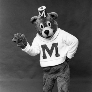Black-and-white historical photograph of a person dressed in a bear mascot costume, standing against a plain backdrop. The bear mascot is wearing a sweater with a large letter "M" on the front and a small hat with the letter "M" on top. The mascot is posed with one hand on its hip and the other raised in a friendly wave, conveying a welcoming and spirited demeanor.