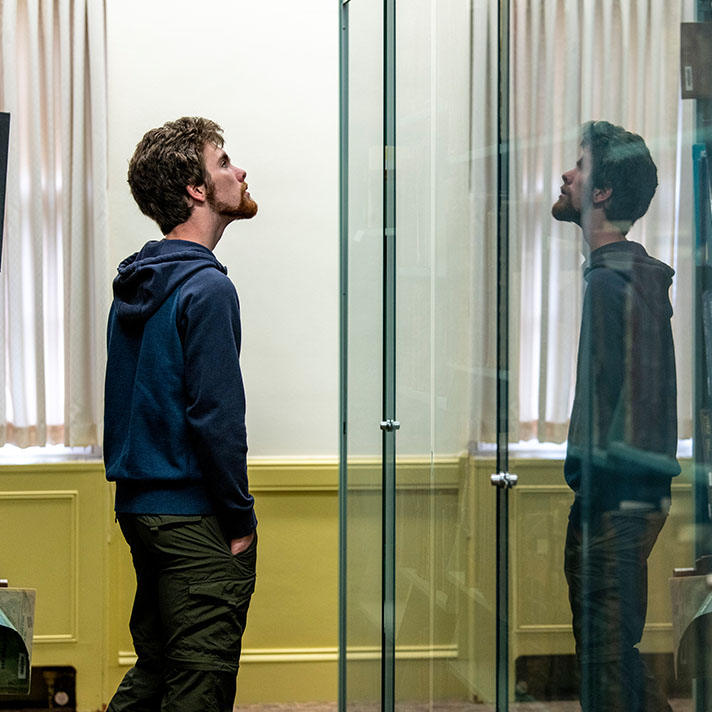 A person wearing a dark hoodie and cargo pants stands in a well-lit room, closely observing a glass display case. Their reflection is visible in the glass, creating a sense of symmetry. The background features tall windows with sheer curtains and pale yellow wainscoting along the walls, contributing to a calm, contemplative atmosphere.