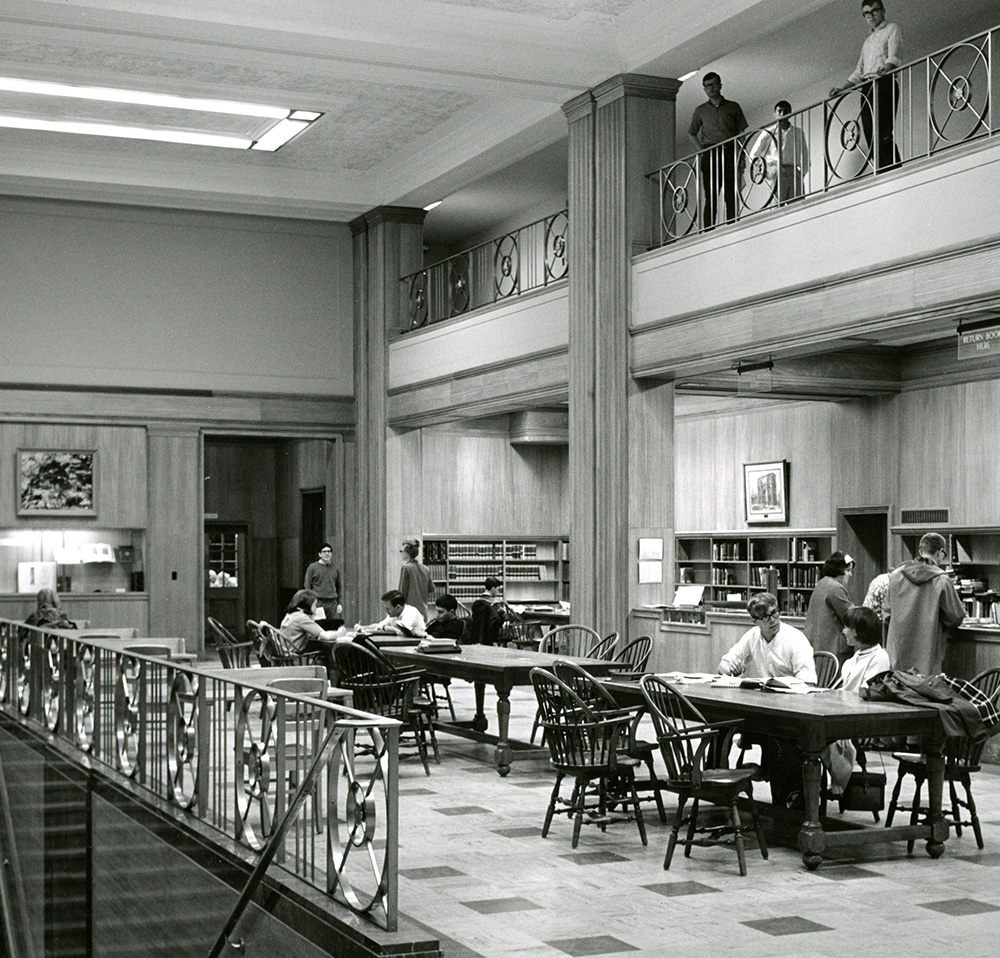 A black and white photograph from 1965 shows the spacious second floor of a library, bustling with students. In the foreground, a decorative metal railing separates the viewer from the main study area. Several students are seated at large wooden tables with chairs, reading and studying. In the background, more students browse bookshelves and interact at a service desk. Above, a balcony level overlooks the main floor, with a few students visible leaning over the railing, observing the activity below. The room features tall pillars, wood-paneled walls, and a patterned floor, with natural light coming from an unseen source and overhead fluorescent lighting illuminating the space.