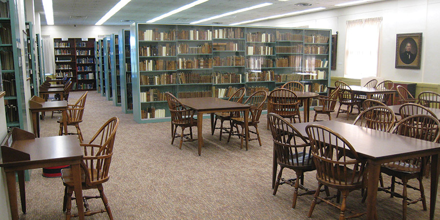 The University of Maine Special Collections Reading Room, featuring rows of wooden tables and chairs arranged for study, glass-fronted bookcases filled with rare books, and a carpeted floor. The room is well-lit by overhead fluorescent lights and natural light from a window. A portrait hangs on the wall, and individual study desks line the left side of the room.