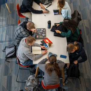 An overhead view of seven students sitting around a rectangular table in a study area with blue carpeted flooring. Some students are using laptops or smartphones, while others are reading or talking. The group appears to be engaged in collaborative work or studying in a casual academic setting.