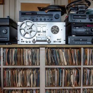 A collection of vintage audio equipment, including a reel-to-reel tape recorder, CD players, and cassette decks, is displayed on top of a large wooden shelf filled with tightly packed vinyl records. The records are organized vertically in compartments, and several blank tapes and discs are stacked on top of the devices. The setting suggests a personal archive or media preservation workspace.
