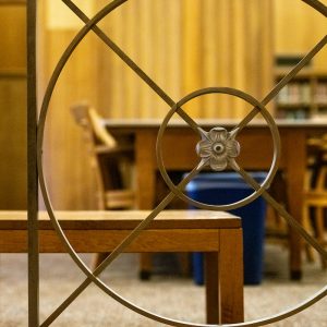 A decorative railing with a circular floral design in the center is in the foreground, partially obscuring a view of a quiet library space. In the background, wooden tables and chairs are arranged for study, with bookshelves and wood-paneled walls adding warmth to the environment.