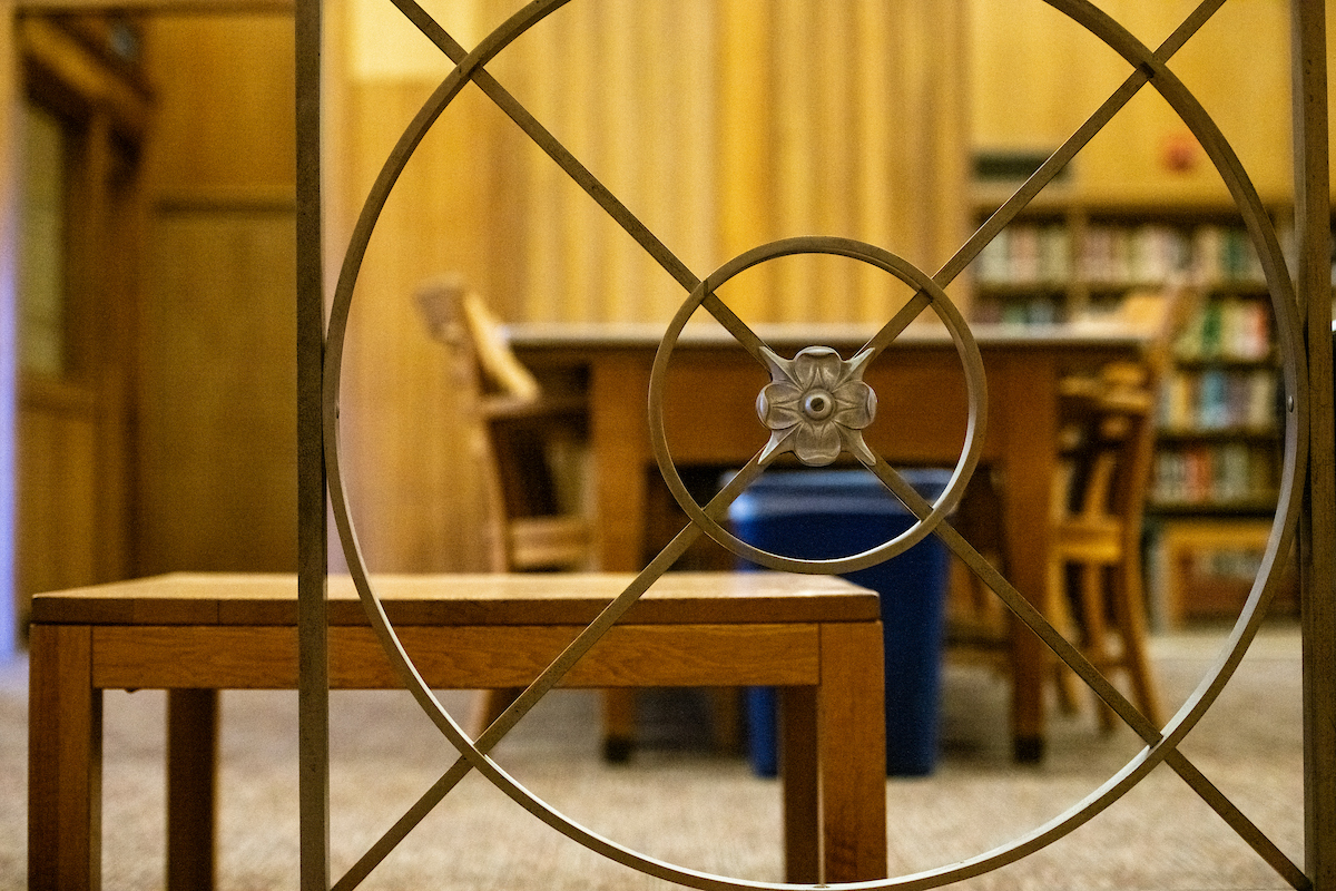 A decorative railing with a circular floral design in the center is in the foreground, partially obscuring a view of a quiet library space. In the background, wooden tables and chairs are arranged for study, with bookshelves and wood-paneled walls adding warmth to the environment.