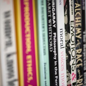 A close-up of book spines on a library or bookstore shelf, featuring titles on topics such as feminist philosophy, ethics, emotions, and race. The titles are tightly packed and vary in color, with some words clearly legible.