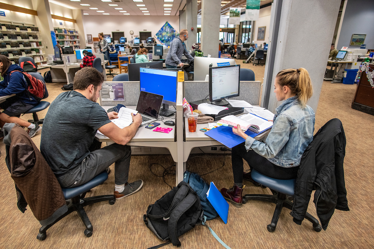 Two students sit across from each other at a shared computer workstation in a busy academic library, studying with laptops, papers, and textbooks open. Other library users are visible in the background at computers and tables under bright lighting.
