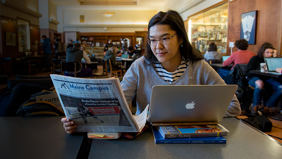 A person with glasses and long dark hair sits at a table in a busy café or study area. They are reading a newspaper titled "Maine Campus" while working on a silver MacBook laptop, which is propped up on two textbooks. They wear a striped shirt under a gray sweater and have a focused expression. In the background, several other people are engaged in conversations or working on laptops, with bookshelves and a display case visible on the wall.