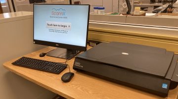A ScannX Book ScanCenter 6000, consisting of a large flatbed scanner and a Dell monitor displaying the 'Touch here to begin' screen, sits on a light wooden desk with a keyboard and mouse.