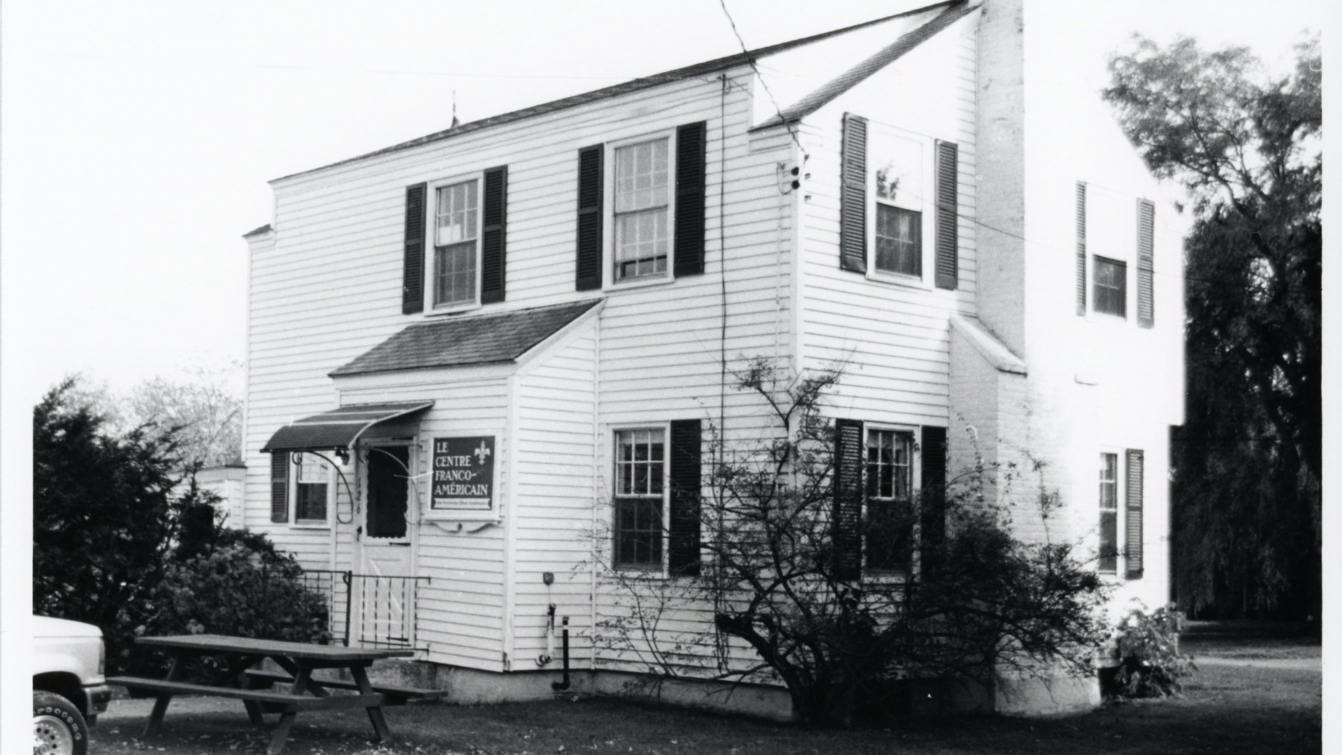 A black and white photograph of a two-story house with white siding and dark shutters on the windows. The house has a small porch with an awning over the entrance. A sign next to the door reads 'Le Centre Franco-Américain' with a cross symbol above it. In front of the house, there is a picnic table and some bushes. A portion of a vehicle is visible on the left side, and trees are in the background.